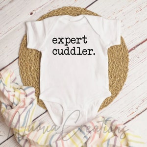 Könnte beinhalten: Weißer Baby-Body mit dem Text "expert cuddler" in Schwarz. Der Body liegt auf einer gewebten Matte, mit einer gestreiften Decke in der Ecke. Der Hintergrund ist helles Holz.
