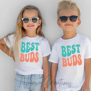 May include: Two children wearing white t-shirts with the text "Best Buds" in turquoise and peach colored letters. The children are wearing sunglasses and smiling.