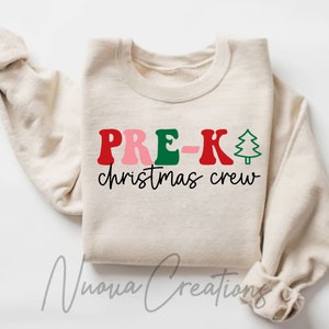 Puede incluir: Sudadera color crema con el texto "PRE-K christmas crew" en rojo, rosa, verde y negro. Un gráfico de árbol de Navidad verde está al lado de la "K". La sudadera tiene mangas largas y cuello redondo.