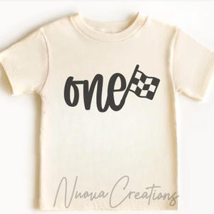 May include: Cream-colored toddler t-shirt with the word "one" in a bold, black script, accompanied by a checkered flag graphic. The shirt has short sleeves and a crew neck. The text "Nuova Creations" is printed at the bottom.