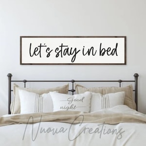May include: A white wooden sign with a black handwritten message that reads "Let's stay in bed". The sign is hanging above a bed with white and beige bedding.