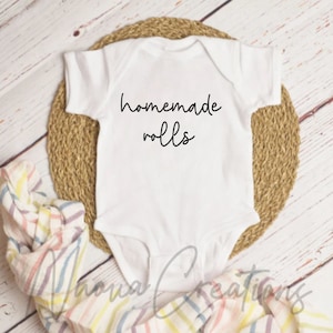 Pode incluir: Um macacão branco para bebê com texto preto que diz "homemade rolls". O macacão está em um tapete de palha trançado.