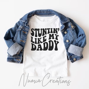 May include: White t-shirt with black text that reads "STUNTIN' LIKE MY DADDY" in a retro font. The shirt is folded and displayed on a denim jacket.
