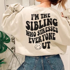 May include: A beige sweatshirt with the text "I'm the sibling who stresses everyone out" in black, with a smiley face with two Xs for eyes and a tongue sticking out.