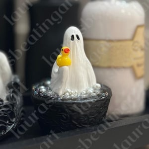 May include: A whimsical Halloween-themed figurine of a ghost holding a yellow rubber duck in a black tub filled with bubbles. The ghost is draped in a white sheet with black eye holes. A decorative bottle is in the background.