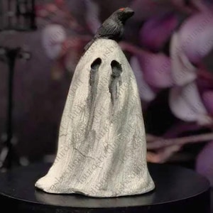 May include: A light grey, textured ghost figurine draped in a sheet-like form with two dark, hollow eye sockets. A small, detailed black crow with a single red eye is perched on the very top of the ghost's head, adding a dark accent to the piece.
