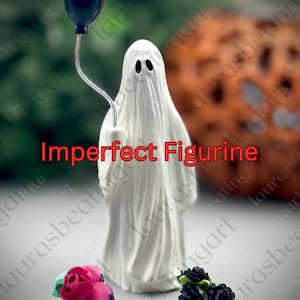 Op de afbeelding: Een wit spookfiguurtje met twee zwarte oogopeningen, dat een dunne draad vasthoudt met een kleine zwarte ballon. De tekst Imperfect Figurine staat in het rood in het midden. Een roze bloem en zwarte bessen bevinden zich aan de onderkant.