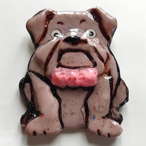 Puede incluir: Una figura artesanal de cerámica o resina brillante de un bulldog con cuerpo de color malva claro, contornos pintados en marrón oscuro y una lengua rosa prominente. El perro tiene una cara ancha y plana, ojos blancos grandes y acabado liso.