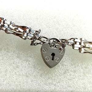 Puede incluir: Una pulsera de metal plateado que presenta una serie de eslabones rectangulares entrelazados y retorcidos. La pulsera incluye un dije en forma de candado de corazón con una cerradura central y cuatro detalles tipo tornillo.