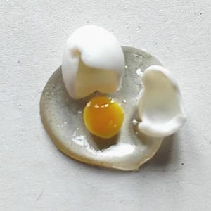 May include: A close-up of a cracked egg on a small, round, grey plate. The egg white partially covers the yolk, which is a bright yellow-orange colour. A piece of the eggshell is visible.