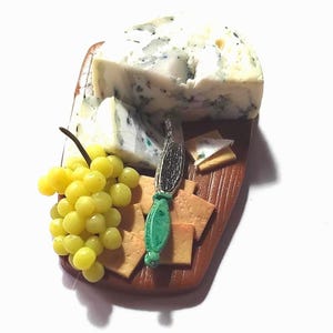 May include: A miniature decorative board featuring a wedge of blue cheese, a cluster of yellow grapes, several square crackers, and a small knife with a green handle. The board has a brown, textured surface resembling wood grain and is arranged.