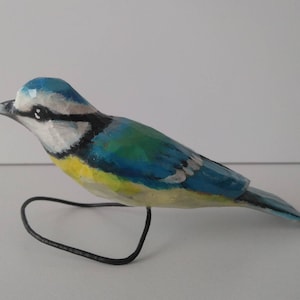Puede incluir: Una figurita de pájaro azul de madera pintada a mano con pintura azul, verde, amarilla y blanca. El pájaro está posado en un soporte de alambre negro.