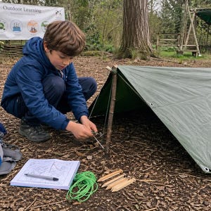 Puede incluir: Una persona con una chaqueta azul está montando un refugio de lona verde en un área boscosa. Hay una mochila, un cuaderno con un mapa, una cuerda verde y estacas de madera cerca. Una pancarta dice "Outdoor Learning".