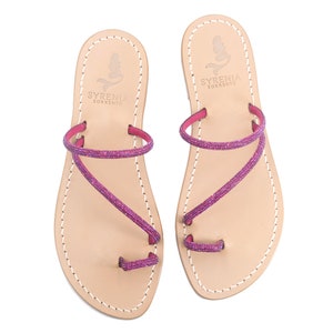 May include: A pair of pink leather sandals with a criss-cross strap design. The straps are adorned with pink glitter. The sandals have a leather sole and are branded with the text "Syrenia Sorrento".