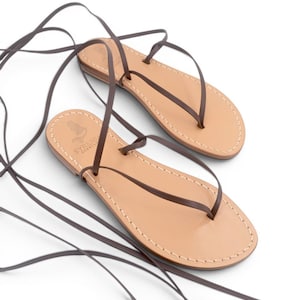 Può includere: Un paio di sandali piatti con solette in pelle color cuoio chiaro, cuciture bianche lungo i bordi e lunghi lacci in pelle marrone scuro da allacciare. La zona del tallone della soletta presenta un logo con la scritta SYRENIA.