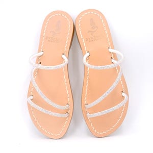 May include: White leather sandals with three straps adorned with rhinestones. The sandals have a tan leather sole and the brand name "Syrenia Sorrento" is printed on the insole.