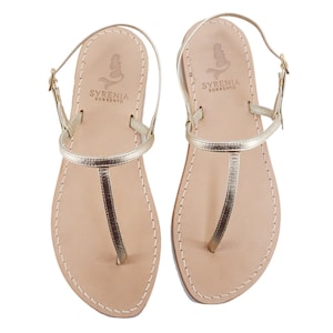 May include: A pair of gold leather sandals with a t-strap design. The sandals have a simple, minimalist style and feature a small mermaid logo on the strap that says "Syrenia Sorrento".