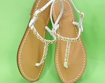 May include: A pair of white leather sandals with gold-colored embellishments. The sandals have a T-strap design with a buckle closure and a light brown sole. The sandals are displayed on a green background.