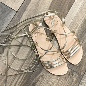 Puede incluir: Sandalias de cuero doradas con cordones y una etiqueta de marca que dice "SYRENIA SOLLENTIO".