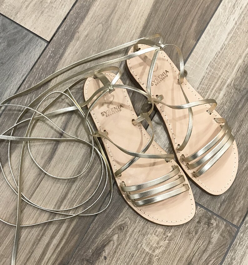 Puede incluir: Sandalias de cuero doradas con cordones y una etiqueta de marca que dice "SYRENIA SOLLENTIO".