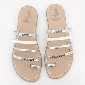 Op de afbeelding: Witte leren sandalen met zilveren en witte banden. De sandalen hebben een teenring en de merknaam "SYRENIA SORRENTO" is op de bovenkant van elke sandaal gedrukt.