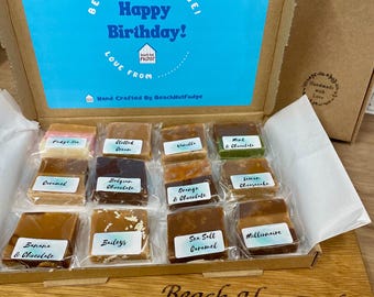 May include: A cardboard gift box containing twelve individually wrapped fudge squares with labels including Fudge Ice, Clotted Cream, Vanilla, Mint & Chocolate, Caramel, Belgian Chocolate, Orange & Chocolate, Lemon Cheesecake, Banana & Chocolate, Baileys, Sea Salt Caramel, and Millionaire. The box lid features the text Beach Hut Fudge! Happy Birthday! Love From.