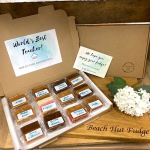 May include: A brown cardboard box with a gift message that says "World's Best Teacher!" and a small card that says "We hope you enjoy your fudge!" The box is filled with small squares of fudge in various flavours. The box is sitting on a wooden chopping board with the words "Beach Hut Fudge" engraved on it.