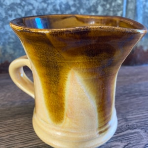 Könnte beinhalten: Eine handgefertigte Keramiktasse mit einer einzigartigen, ausgestellten Form. Die Tasse hat eine warme Farbpalette aus Hellgelb und Dunkelbraun mit glänzendem Finish. Sie hat einen kleinen Henkel und ist für heiße oder kalte Getränke geeignet.