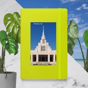May include: A bright yellow notebook with a rectangular image of a white building with a tall spire against a blue sky. The text "Phoenix, AZ" is above the image. A green elastic band secures the notebook.