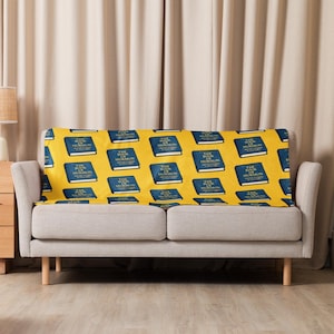 May include: A yellow blanket with a repeating pattern of blue books titled "The Book of Mormon" is draped over a gray sofa. The blanket's design features the book's title in white lettering.
