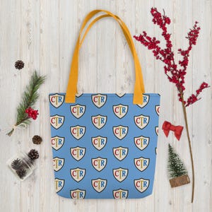 May include: A light blue tote bag with a repeating pattern of shields. Each shield has the letters "CTR" in a gold color. The bag has yellow handles. The bag is on a light wood surface with holiday decorations.