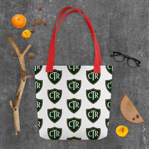 May include: White tote bag with a repeating pattern of green and gold crests with the letters "CTR". The bag has red handles. The bag is on a gray surface.