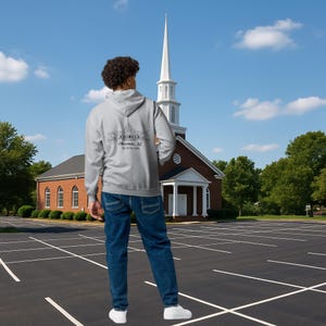 May include: A gray hoodie with a drawing of a building and the text "Phoenix, AZ" on the back. The person is wearing blue jeans and white sneakers. A church building is in the background.