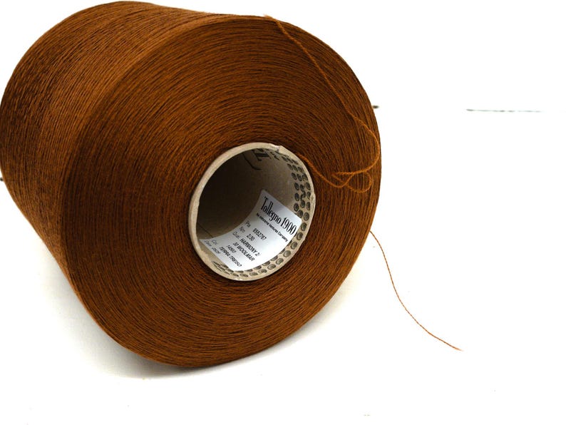 May include: A large spool of brown yarn, ideal for knitting or crochet projects. The yarn is tightly wound and has a smooth texture. The label on the spool reads "Tollegno 1900".