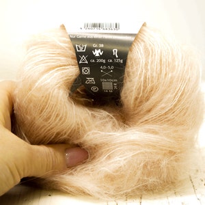 K&ouml;nnte beinhalten: Eine Kn&auml;uel hellrosa Garn mit einem schwarzen Etikett, das Pflegehinweise und Details wie "Gr. 38", "ca. 200g", "ca. 125g", "4.0-5.0", "10x10cm" und "16 M/M" enth&auml;lt.