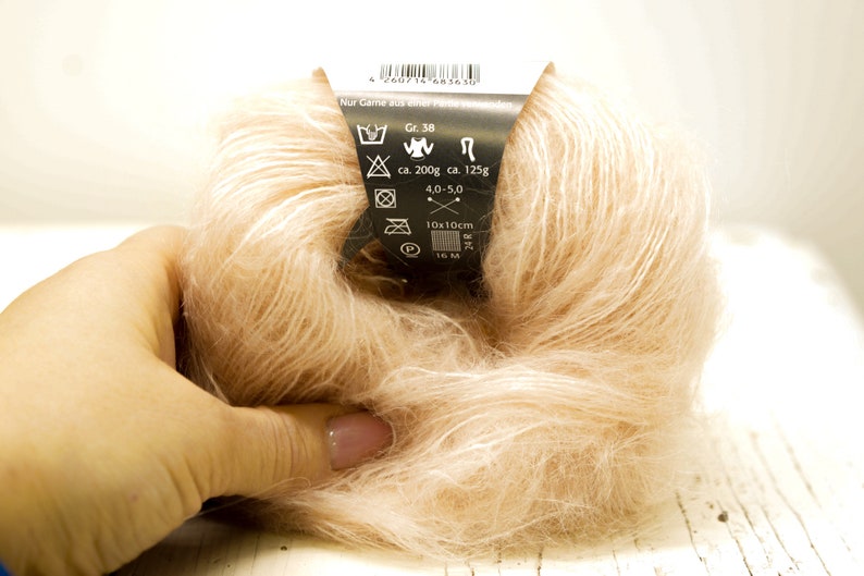K&ouml;nnte beinhalten: Eine Kn&auml;uel hellrosa Garn mit einem schwarzen Etikett, das Pflegehinweise und Details wie "Gr. 38", "ca. 200g", "ca. 125g", "4.0-5.0", "10x10cm" und "16 M/M" enth&auml;lt.