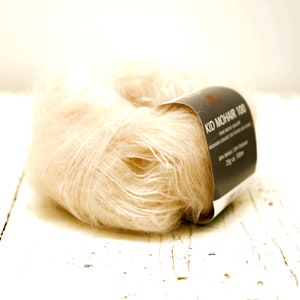 K&ouml;nnte beinhalten: Eine Spule weiches, hellbeiges Garn mit der Aufschrift "Kid Mohair 100".