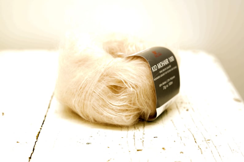 K&ouml;nnte beinhalten: Eine Spule weiches, hellbeiges Garn mit der Aufschrift "Kid Mohair 100".