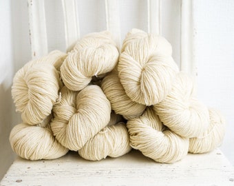 May include: A pile of off-white yarn balls, arranged in a pyramid shape, on a white wooden surface.