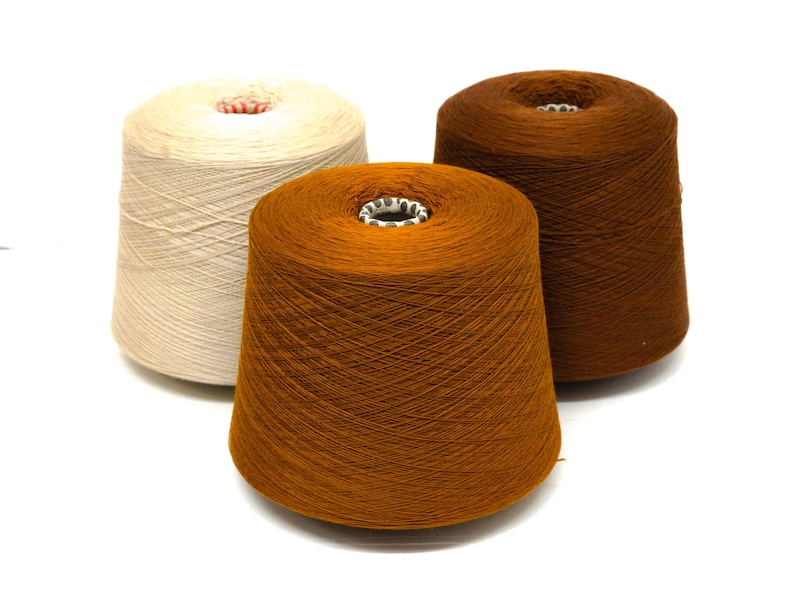 May include: Three spools of yarn in varying shades of brown and cream sit against a white background. The yarn is tightly wound around a central core. The colors range from a light cream to a deep, rich brown, suitable for knitting or crochet projects.