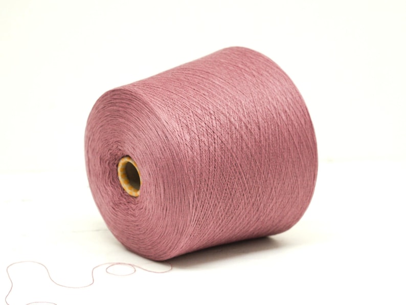 May include: A large spool of dusty rose-colored yarn. The yarn is tightly wound around a cardboard core, with a strand extending from the spool. Ideal for knitting, crocheting, or other fiber arts projects.