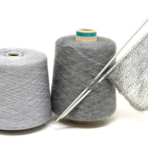 Puede incluir: Dos carretes de hilo gris con una aguja de tejer y una pequeña muestra de tejido de punto gris.