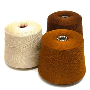 May include: Three cones of yarn in shades of beige, brown, and orange. The yarn is tightly wound around a central cardboard core. The cones are arranged on a white surface, with the yarn fibers visible.