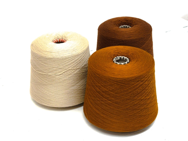 May include: Three cones of yarn in shades of beige, brown, and orange. The yarn is tightly wound around a central cardboard core. The cones are arranged on a white surface, with the yarn fibers visible.