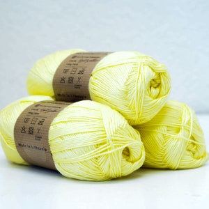 Può includere: Tre gomitoli di filato giallo con etichette marroni che includono istruzioni di lavaggio e il testo "Made in Lithuania".