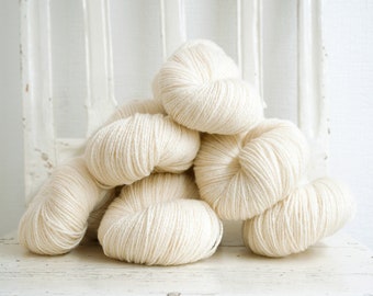 May include: A pile of six skeins of off-white yarn, arranged in a pyramid shape on a white surface.