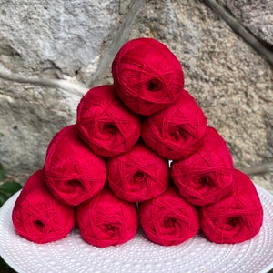 May include: A pile of ten red yarn balls stacked in a pyramid shape on a white plate. The yarn balls are arranged in a symmetrical pattern.