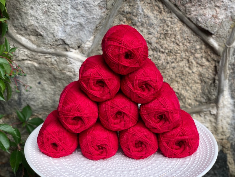 May include: A pile of ten red yarn balls stacked in a pyramid shape on a white plate. The yarn balls are arranged in a symmetrical pattern.