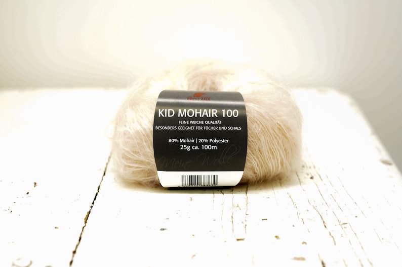 K&ouml;nnte beinhalten: Eine Kn&auml;uel Kid Mohair 100 Garn in einer hellbeigen Farbe. Das Etikett lautet "Kid Mohair 100", "Feine Weiche Qualitat", "Besonders Geeignet Fur Tucher Und Schals", "80% Mohair | 20% Polyester", "25g ca. 100m".