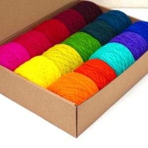 Op de afbeelding: Een kartonnen doos gevuld met kleurrijke garenklossen in een regenboogvolgorde, beginnend met roze en eindigend met blauw.